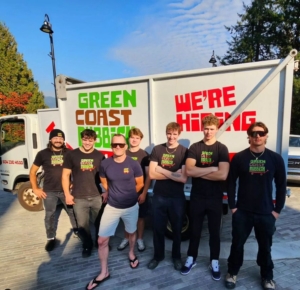 A group of seven people stand smiling in front of a white truck with signs reading GREEN COAST RUBBISH and WERE HIRING in bold letters. Trees and a clear blue sky are visible in the background.